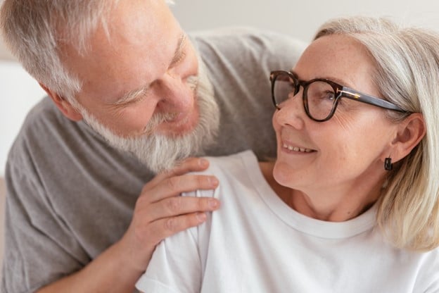 A middle aged man with gray hair, wearing a gray t shirt, embraces a woman from behind with his hand on her shoulder