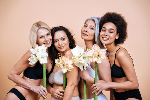 diverse women of different ages pose together in neutral toned lingerie, holding large, vibrant amaryllis flowers