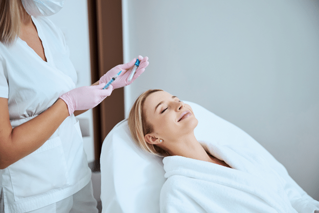 A healthcare professional, wearing a white uniform, pink gloves, and a face mask, is holding a syringe and a small vial, preparing to inject platelet rich plasma