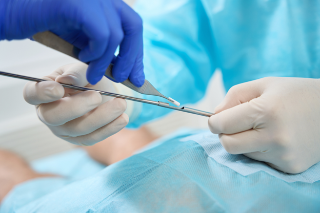A healthcare professional wearing blue gloves uses surgical instruments to handle a small white hormone pellet during a medical procedure on a draped patient